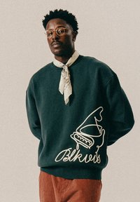 Dark green sweatshirt featuring a white graphic design and text "Blkvis" on the front. The model wears a patterned scarf around the neck.