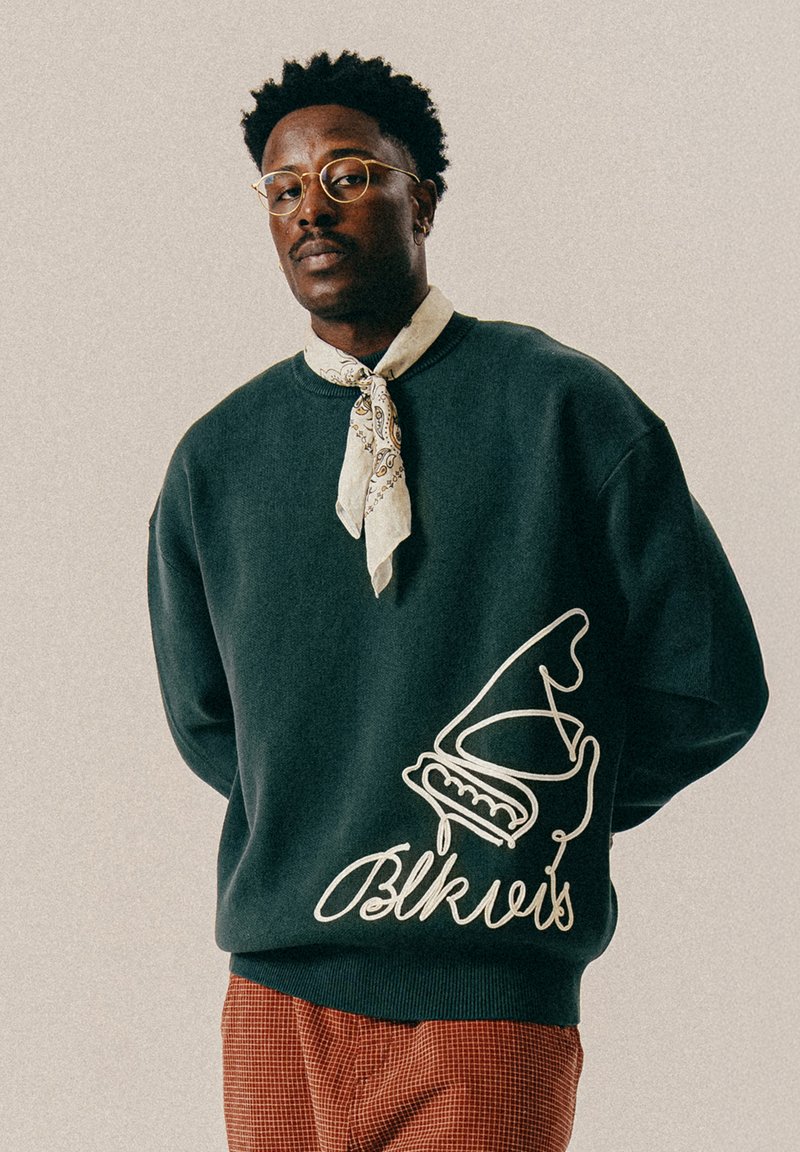 Dark green sweatshirt featuring a white graphic design and text "Blkvis" on the front. The model wears a patterned scarf around the neck.