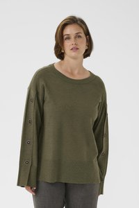 Mujer con suéter verde oliva con detalles de botones a lo largo de las mangas y pantalones grises, de pie contra un fondo claro y sencillo.