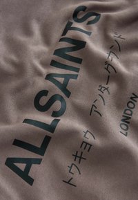 Tessuto beige con una superficie liscia che presenta grande testo verde scuro "ALLSAINTS" e caratteri giapponesi, insieme a "LONDON" in un testo più piccolo.