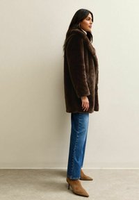 Bruine nepleren jas met lange mouwen en een open voorkant, gecombineerd met blauwe rechte jeans en bruine puntige hakken. Gladde textuur.