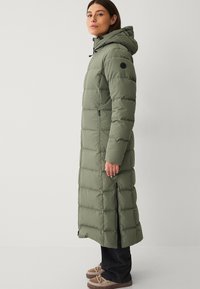 Giacca di piumino verde oliva con cappuccio, design lungo, texture trapuntata, due zip laterali e un logo circolare sulla manica sinistra.