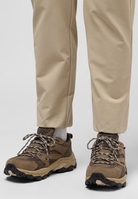 Brown hiking shoes with mesh and suede materials, featuring a rugged black sole and reflective, multi-colored laces. Beige pants visible.