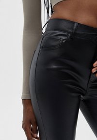 Pantalon noir en simili cuir avec une taille haute, des poches latérales et une coupe ajustée. La surface est lisse avec un léger éclat, mettant en valeur une couture nette.