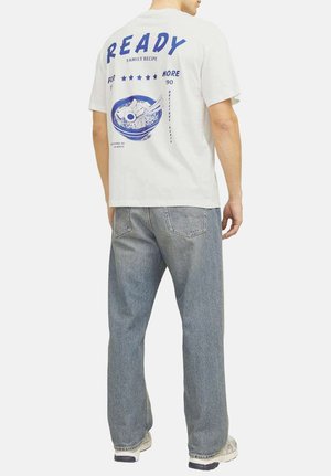 T-shirt blanc à manches courtes avec des graphiques bleus au dos, représentant un bol de nouilles et le texte "RECETTE DE FAMILLE PRÊTE". Porté avec un jean gris.