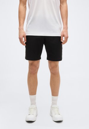 Άνδρας που φοράει μαύρο σορτς, λευκές κάλτσες, λευκά αθλητικά παπούτσια και λευκό κοντομάνικο πουκάμισο, εμφανιζόμενος από το μέσο του μηρού έως τους ώμους σε λευκό φόντο.