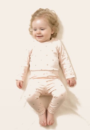 Smilende småbarn med krøllet blondt hår, der ligger ned, iført lyserødt pyjamas med små hjerteprint.