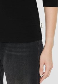 Schwarzes T-Shirt mit figurbetontem Schnitt, kurzen Ärmeln und einem Etikett an der Seitennaht. Kombiniert mit schwarzen Jeans, die eine dezente Textur aufweisen.