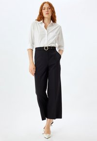 Chemise blanche à boutons, associée à un pantalon noir à taille haute et jambes larges avec une ceinture agrémentée de perles, complétée par des talons aiguilles blancs.