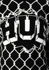 Sort sweater med hvid kæde-link mønster og fed "HUL" tekst. Struktureret strikstof, fremtrædende kontraster i designet, strukturerede former.
