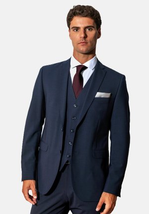 Jonge man die een donkerblauw driedelig pak draagt, een wit overhemd, een bordeauxrode stropdas en een wit pochet, staand tegen een effen lichte achtergrond.