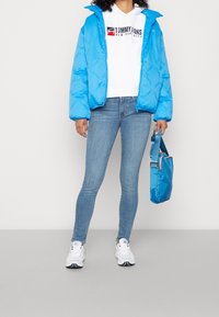 Blue puffer jacket with logo, white hoodie with branding, fitted light blue jeans, and white sneakers. Holding a blue bag.