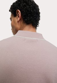 Lyserød strikket poloshirt med en tekstureret overflade, der har en lille krave og en tynd kontrastdetalje. Set fra bagsiden.