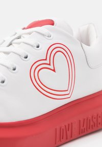 Baskets blanches avec semelle rouge, cœur rouge double contour sur le côté, lacets blancs, et "LOVE MOSCHINO" embossé sur la semelle.