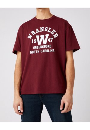 Mężczyzna w bordowej koszulce Wrangler z białym napisem "WRANGLER 1947 Greensboro North Carolina" i ciemnych dżinsach na jednolitym tle.