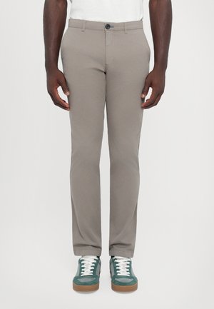 Lys beige bukser i slim fit design, glat tekstur, forlommer og lukning med en enkelt knap. Parret med grønne og hvide sneakers.