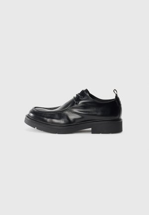 Black leather dress shoe with rounded toe, lace-up front, and a decorative seam. Features a thick, textured rubber sole and a heel tab.