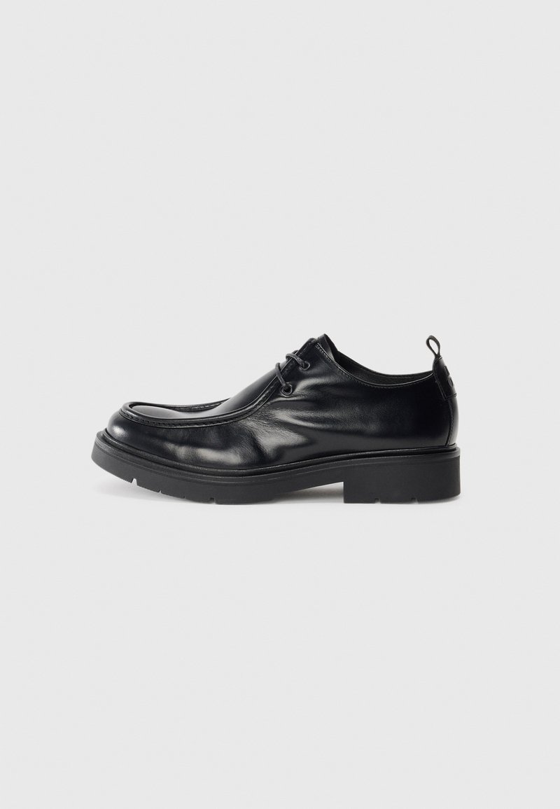 Zapato de vestir de cuero negro con puntera redondeada, parte frontal con cordones y una costura decorativa. Presenta una suela de goma gruesa y texturizada y una pestaña en el talón.