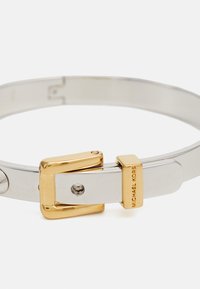 Pulsera bangle de metal en tonos plata y dorado con un diseño de hebilla y grabado "MICHAEL KORS" en una pieza de acento. Textura de superficie lisa.