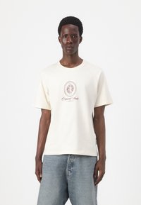 Cremefarvet bomulds t-shirt med korte ærmer, prydet med et centralt grafisk design i bordeaux og detaljer ved halsudskæringen. Bæres med lyseblå jeans.