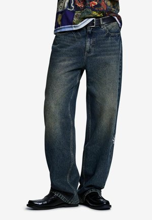 Personne portant un jean bleu foncé ample, une ceinture noire, une chemise à motif floral, et des chaussures de ville noires brillantes sur fond blanc.