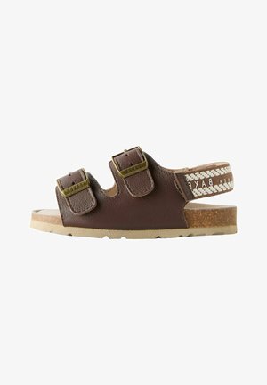Hnědá kožená sandála s dvěma nastavitelnými pásky na přezku, korkovou podrážkou a podpatkem z vzorované látky s textem značky.