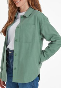 Green button-up shirt made of soft fabric, featuring a chest pocket, rounded hem, and casual fit, paired with blue jeans.
