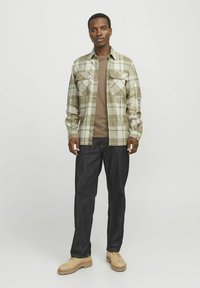 Geruit flanellen shirt in gedempte groentinten, gecombineerd met een bruine t-shirt en donkere jeans. Schoeisel is lichtbruin met een robuuste zool.