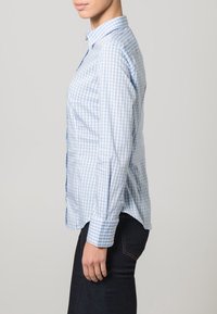Ljust blå rutig skjorta med knappar, krage, långa ärmar och rundad nederkant. Har enkla manschettknappar och en slät tygstruktur.
