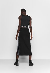 Černý přiléhavý top bez rukávů spojený s dlouhou černou sukní s bílým vzorovaným páskem po straně. Modelka má na sobě černé boty a ponožky.