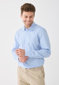 Svetlo modrá košeľa s dlhým rukávom a zapínaním na gombíky, so špicatým golierom a bielymi gombíkmi, kombinovaná s béžovými nohavicami. Hladká textúra, strih prispôsobený postave.