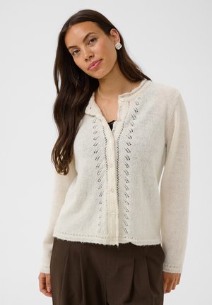 Vrouw met lang bruin haar draagt een wit gebreid vest met kanten details en een bruine broek, kijkt recht vooruit tegen een effen achtergrond.