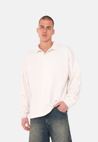 Lys rosa langærmet polo med afslappet pasform, knapkrave og broderet detalje på venstre bryst, kombineret med denimshorts.