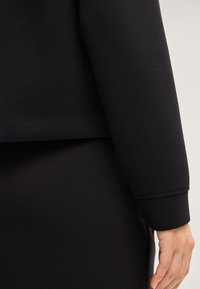 Sweatshirt noir avec une coupe décontractée, en tissu doux, à manches longues et ourlet arrondi. Le poignet a un détail cousu.