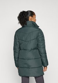 Abrigo acolchado de un verde oscuro con un alto cuello. Presenta costuras en forma de chevron y mangas largas, terminando a mitad del muslo con un dobladillo recto.