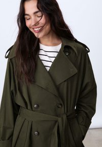 Olivengrøn trenchcoat med dobbeltradet knaplukning, bred krave og bindebælte. Båret over en hvidstribet skjorte. Blød, glat tekstur.