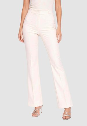 Donna che indossa pantaloni a zampa a vita alta color crema e sandali argento con tacco a spillo e cinturini, in piedi davanti a uno sfondo semplice.