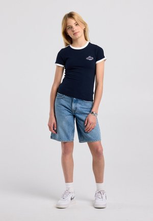 Adolescente debout, portant un t-shirt bleu marine avec bordure blanche, un short en jean, des chaussettes blanches, des baskets blanches et un bracelet argenté au poignet gauche.