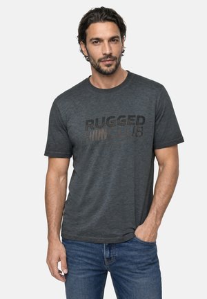 Homme portant un t-shirt "Rugged Club" gris foncé et un jean bleu, posant avec une main dans la poche devant un fond uni.