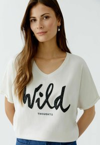 Lässiges, weißes T-Shirt mit V-Ausschnitt, das ein großes, glitzerndes schwarzes "Wild"-Textdesign und das Wort "THOUGHTS" darunter zeigt.