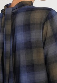 Chemise à carreaux avec un schéma de couleurs bleu marine et vert olive, col montant et détail noué dans le dos. Texture de tissu léger.