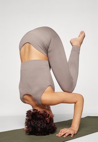 Ribbed, brown activewear set features a high-waisted bottom and cutout design. Subject is in a headstand on a yoga mat.