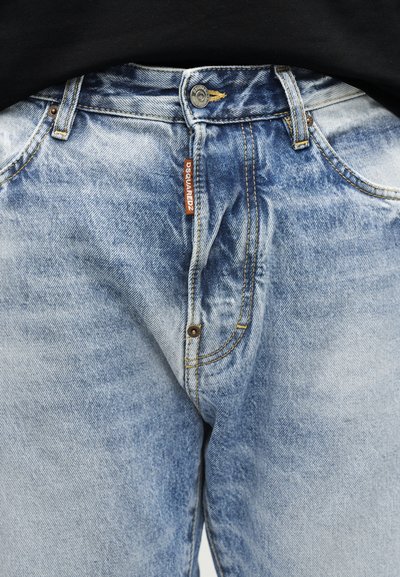 Jean bleu clair délavé avec une étiquette de marque DSQUARED2 visible près de la fermeture par bouton et des passants de ceinture, porté avec un haut noir.