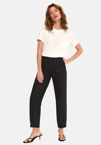 Biała koszulka z krótkim rękawem i kieszenią, w połączeniu z czarnymi spodniami o wysokim stanie. Modelka ma na sobie czarne sandały z złotym akcentem.