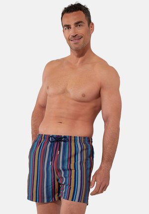 Hemdloser Mann mit heller Haut und dunklem Haar, der bunte, vertikal gestreifte Badeshorts mit Kordel trägt, lächelt und vor einem weißen Hintergrund posiert.