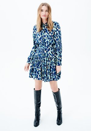 Abito a maniche corte con un motivo blu, verde e bianco, e gonna plissettata. Abbinato a stivali neri in pelle al ginocchio.