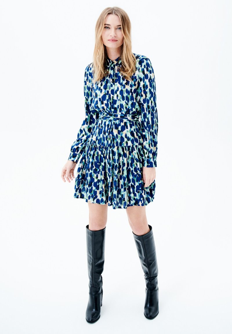 Vestido de manga corta en un patrón azul, verde y blanco, con falda plisada. Combinado con botas de cuero negro hasta la rodilla.