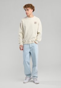 Sudadera de cuello redondo beige con un pequeño gráfico marrón, combinada con unos jeans sueltos de color azul claro y zapatillas blancas. Diseño sencillo y textura suave.