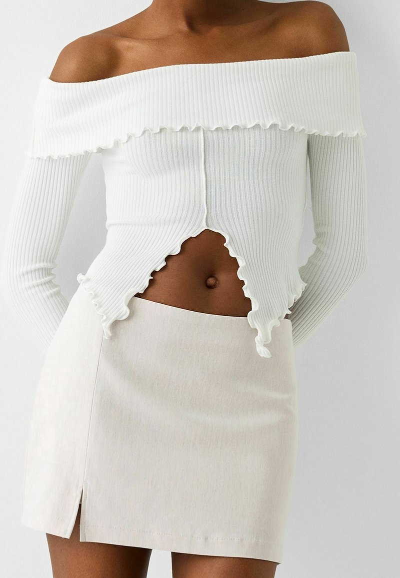 Mörkhyad person som bär en vit off-shoulder ribbad topp med vågiga kanter och en vit miniskjol med en sidofåra.