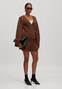 Bruin fuzzy wikkeltrui gecombineerd met een bijpassende flared rok; geaccessoireerd met een zwarte clutch en puntige zwarte hakken.